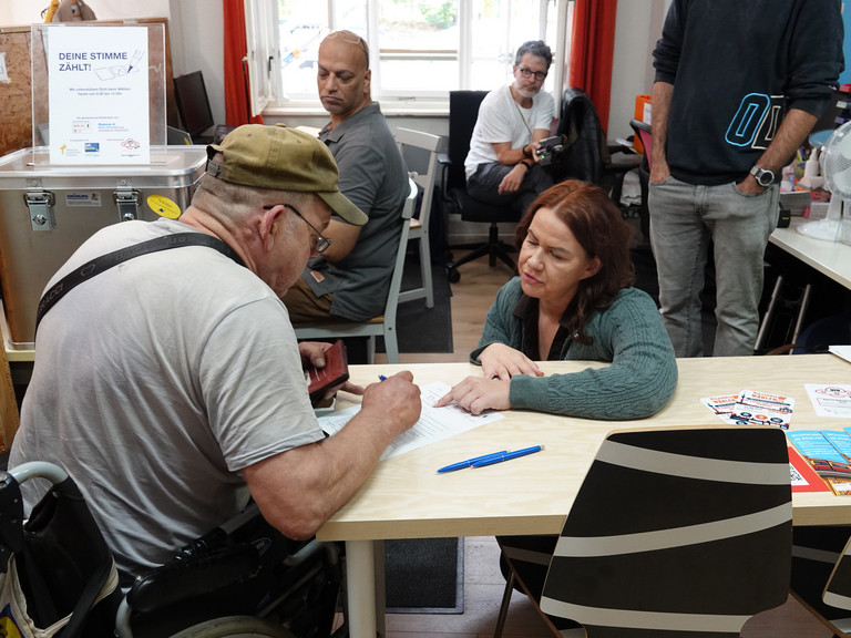 Frau hilft einem Mann beim Eintragen in das Wahlverzeichnis und beim Beantragen der Briefwahlunterlagen in der Tagesstätte des Sozialprojekts Prenzlauer Berg von Immanuel Beratung Frau hilft einem Mann beim Eintragen in das Wahlverzeichnis und beim Beantragen der Briefwahlunterlagen in der Tagesstätte des Sozialprojekts Prenzlauer Berg von Immanuel Beratung